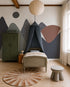 A cozy kids’ bedroom features a single bed beneath a dark canopy, round ceiling light, green locker wardrobe, stacked blankets, sunburst rug, and the Mountain Mural as a charming focal point for nature-inspired décor.