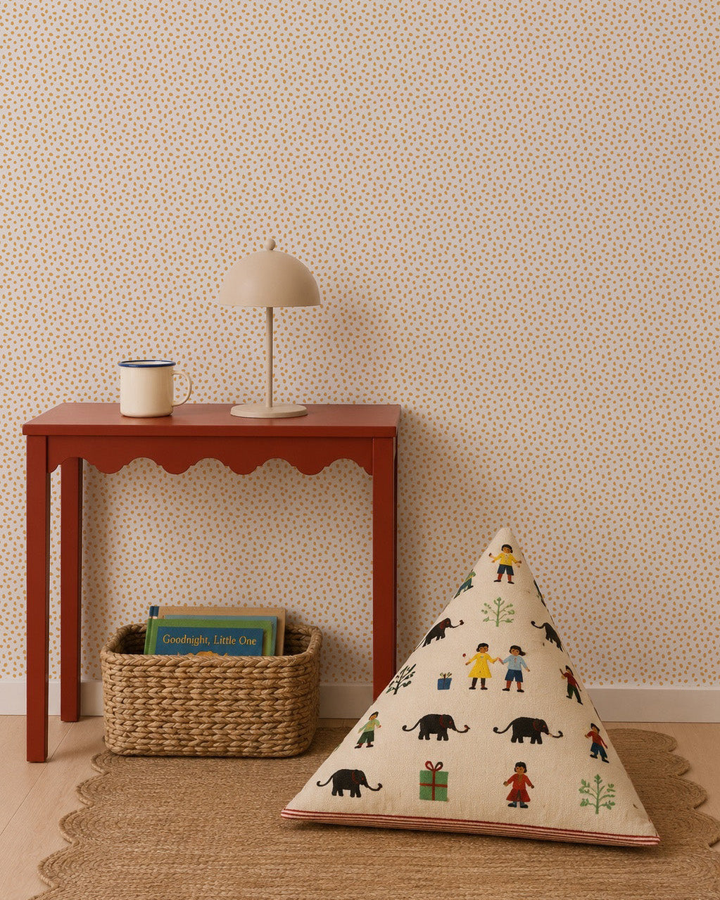 A red table holds a lamp and mug next to a woven basket with books. A triangular cushion featuring colorful people and animal designs sits on a round rug against a statement wall adorned with the Sprinkle Wallpaper in Mustard.