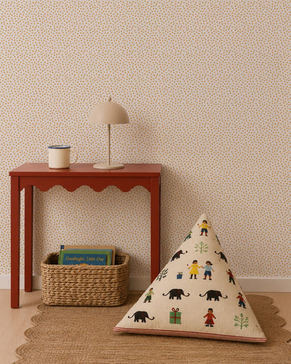 A red table holds a lamp and mug next to a woven basket with books. A triangular cushion featuring colorful people and animal designs sits on a round rug against a statement wall adorned with the Sprinkle Wallpaper in Mustard.