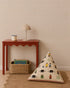 A red table holds a lamp and mug next to a woven basket with books. A triangular cushion featuring colorful people and animal designs sits on a round rug against a statement wall adorned with the Sprinkle Wallpaper in Mustard.