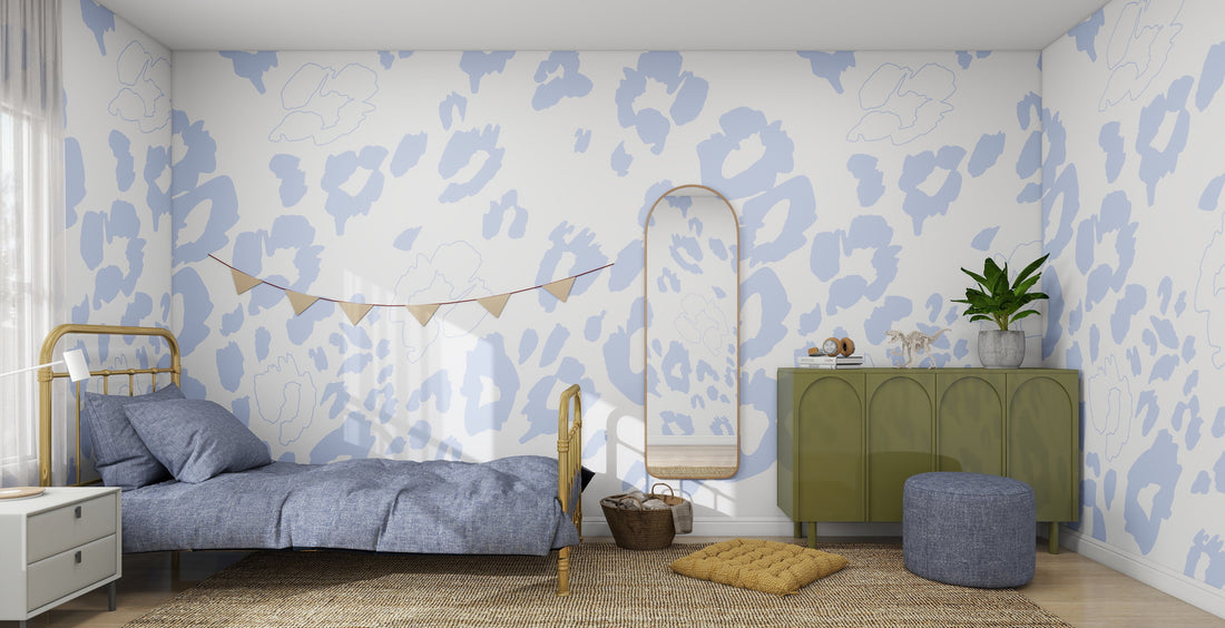 A cozy bedroom features a single bed, green dresser, mirror, ottoman, plants, woven rug on wood floors, and window. The Nelly Leopard Print Mural stands out as the highlight behind the bed, which is adorned with a garland.