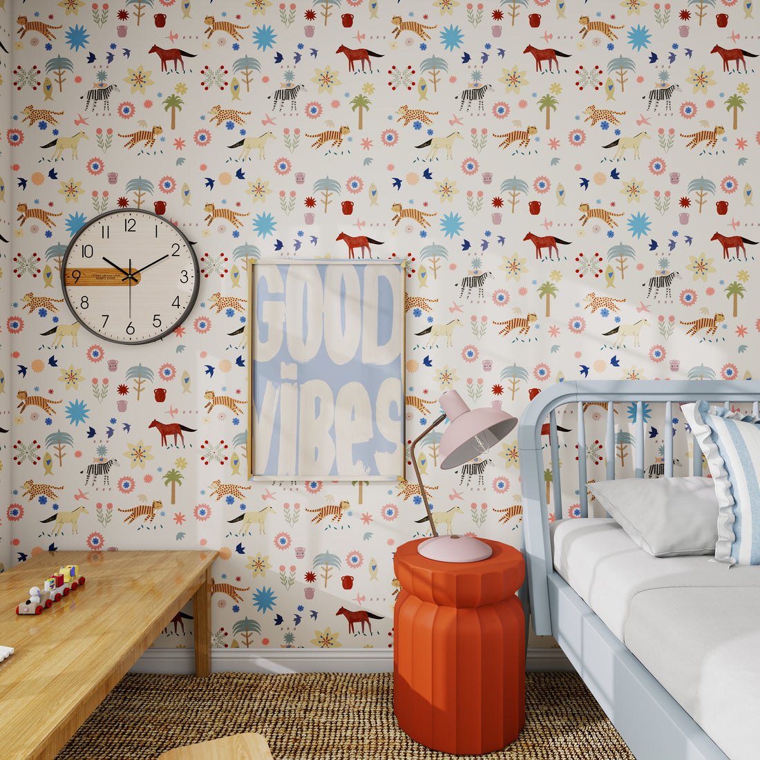 A colorful kids’ room features Little Wanderers Wallpaper, a round wall clock, GOOD VIBES poster, wooden toy table, orange stool, pink lamp, and a white bed with blue bedding to brighten the nursery.