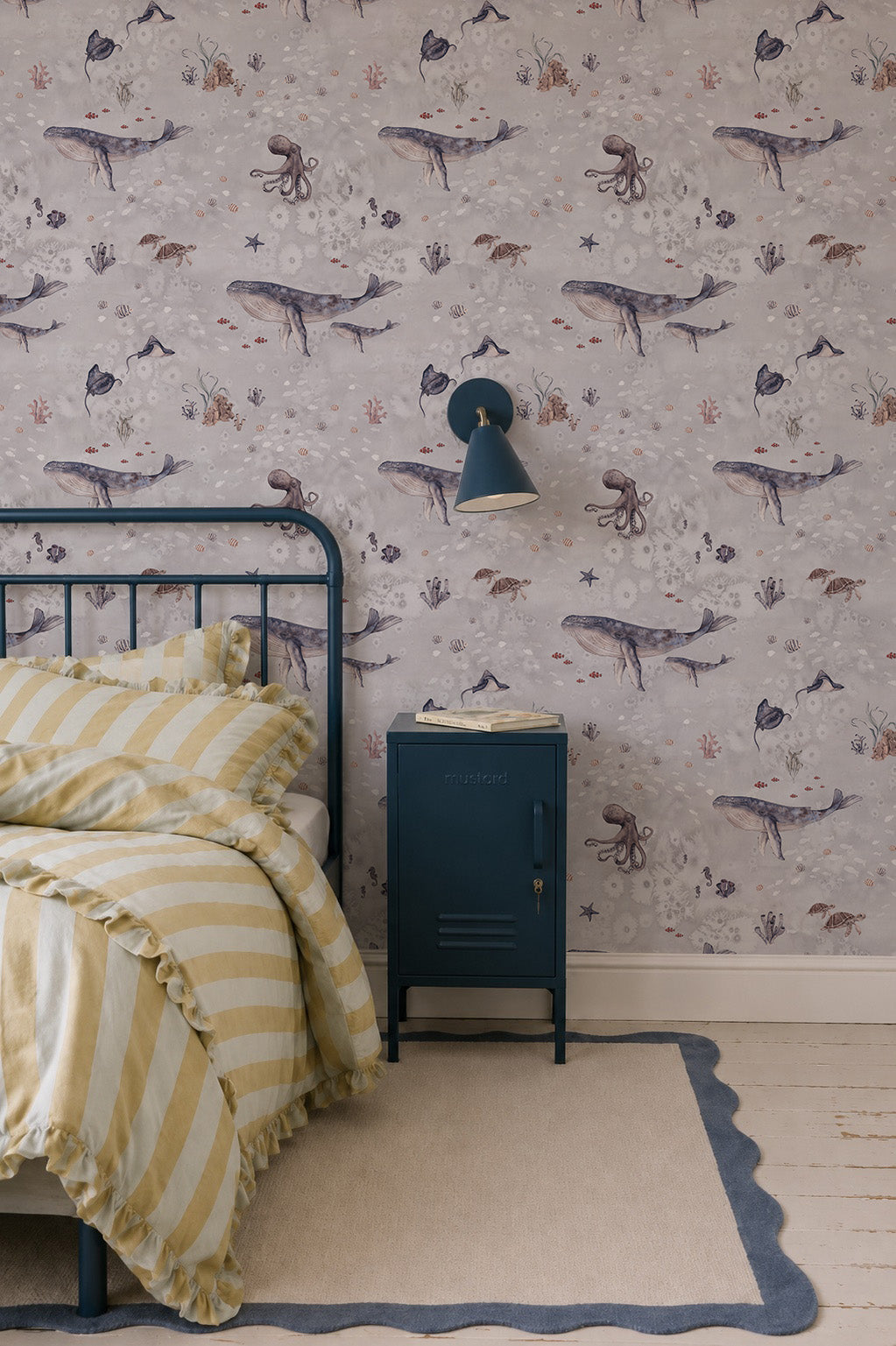 A cozy kids’ room features a blue metal bed, yellow-striped bedding, a navy nightstand and lamp. Above a light rug with navy trim, the Ocean Friends Wallpaper showcases whales and sea creatures for playful decor.