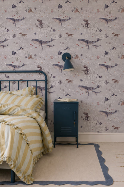 A cozy kids’ room features a blue metal bed, yellow-striped bedding, a navy nightstand and lamp. Above a light rug with navy trim, the Ocean Friends Wallpaper showcases whales and sea creatures for playful decor.