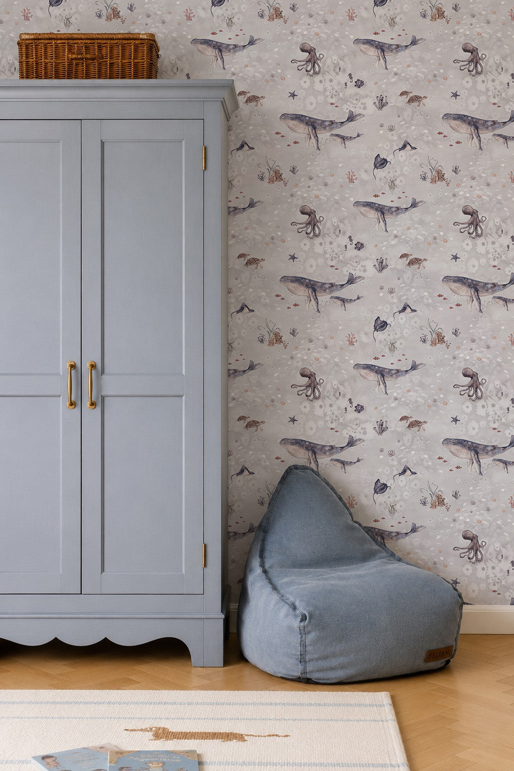 A light blue wardrobe stands next to a blue beanbag chair against the whimsical Ocean Friends Wallpaper in a children’s room, while a wicker basket atop the wardrobe adds to the charming decor.