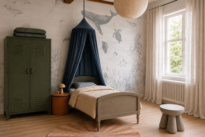 A cozy kids bedroom featuring the eye-catching Ocean Magic Mural in Blue, a green locker wardrobe, a bed with a navy canopy, a side table, a lamp, a round rug, a stool, and a large window with white curtains.