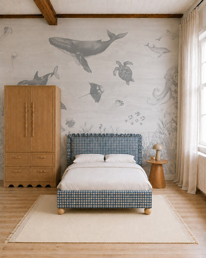 A cozy bedroom with a plaid bed, light rug, wooden furniture, and the Ocean Magic Mural, Blue featuring sea animals like whales and turtles on the wall. Natural light streams in through sheer curtains.