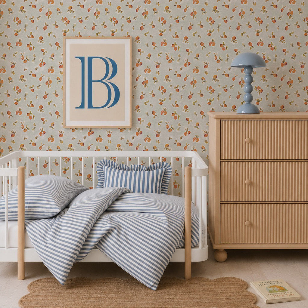 A cozy children’s bedroom with a small white bed, blue-striped bedding, round rug, wooden dresser with a blue lamp, and Ollies Oranges Wallpaper in Blue. A framed print featuring a large blue letter “B” hangs on the wall.
