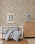 A cozy children’s bedroom with a small white bed, blue-striped bedding, round rug, wooden dresser with a blue lamp, and Ollies Oranges Wallpaper in Blue. A framed print featuring a large blue letter “B” hangs on the wall.