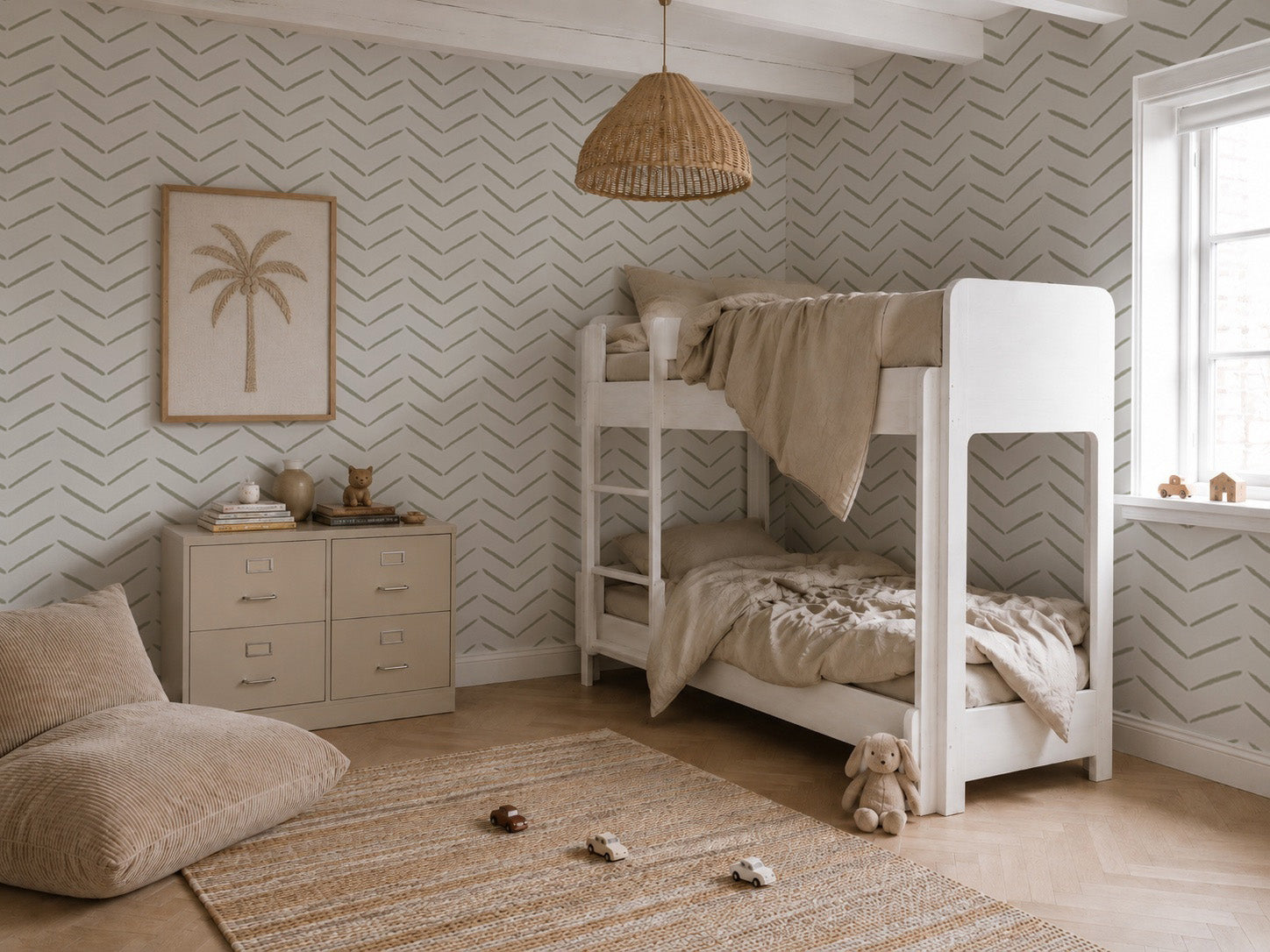 A cozy children’s bedroom features white bunk beds, beige bedding, a chest of drawers, a palm tree picture, and stylish wall decor such as Herringbone Wallpaper in Olive. A woven rug, soft floor cushion, and a window letting in natural light complete the space. Neutral tones and minimalist decor are featured throughout.