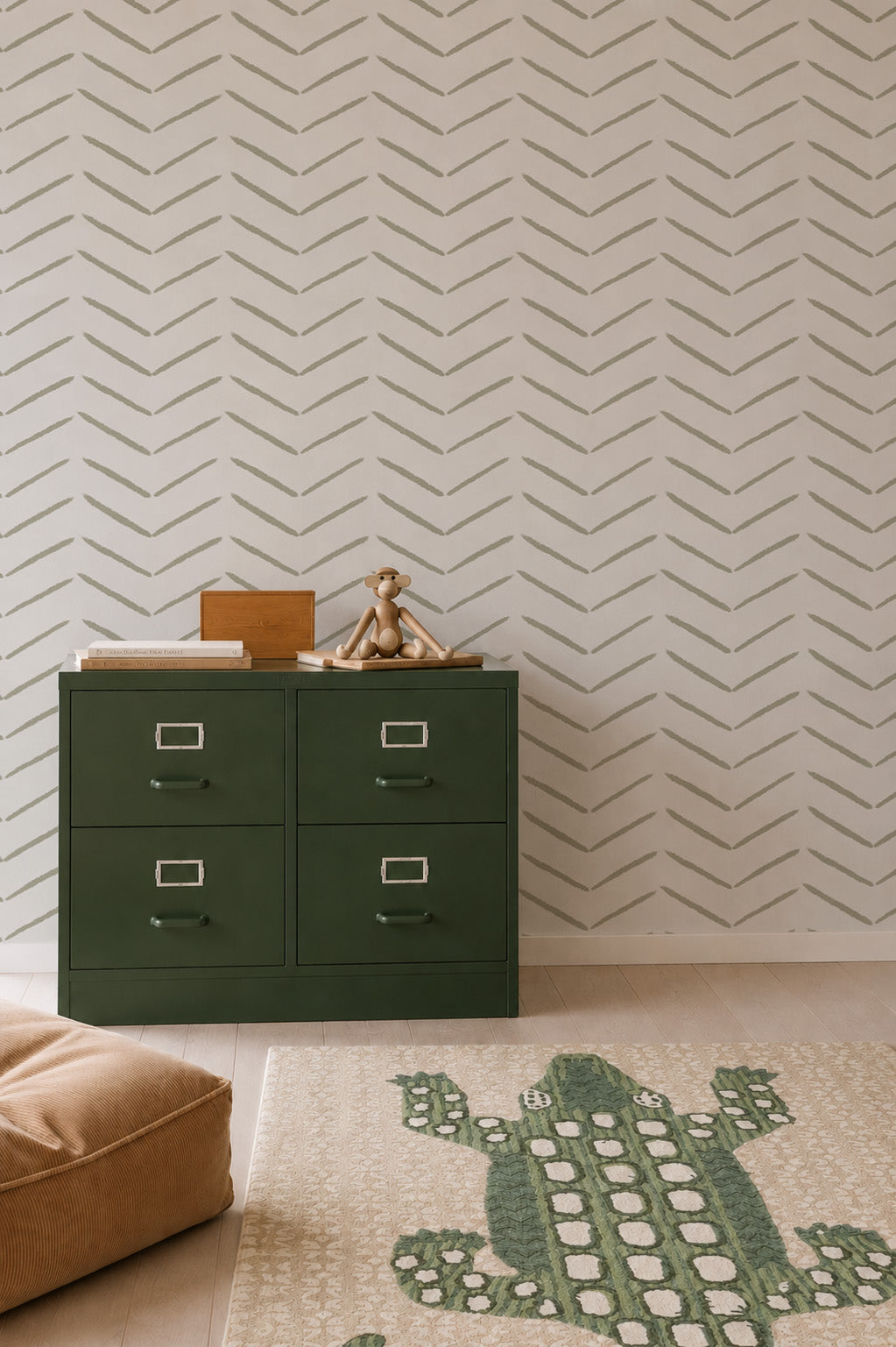 A green drawer cabinet sits against olive herringbone wallpaper, with a toy monkey and boxes on top. A crocodile-patterned rug and tan cushion rest on a light wood floor, creating a space enhanced by stylish wall decor.