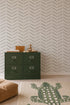A green drawer cabinet sits against olive herringbone wallpaper, with a toy monkey and boxes on top. A crocodile-patterned rug and tan cushion rest on a light wood floor, creating a space enhanced by stylish wall decor.