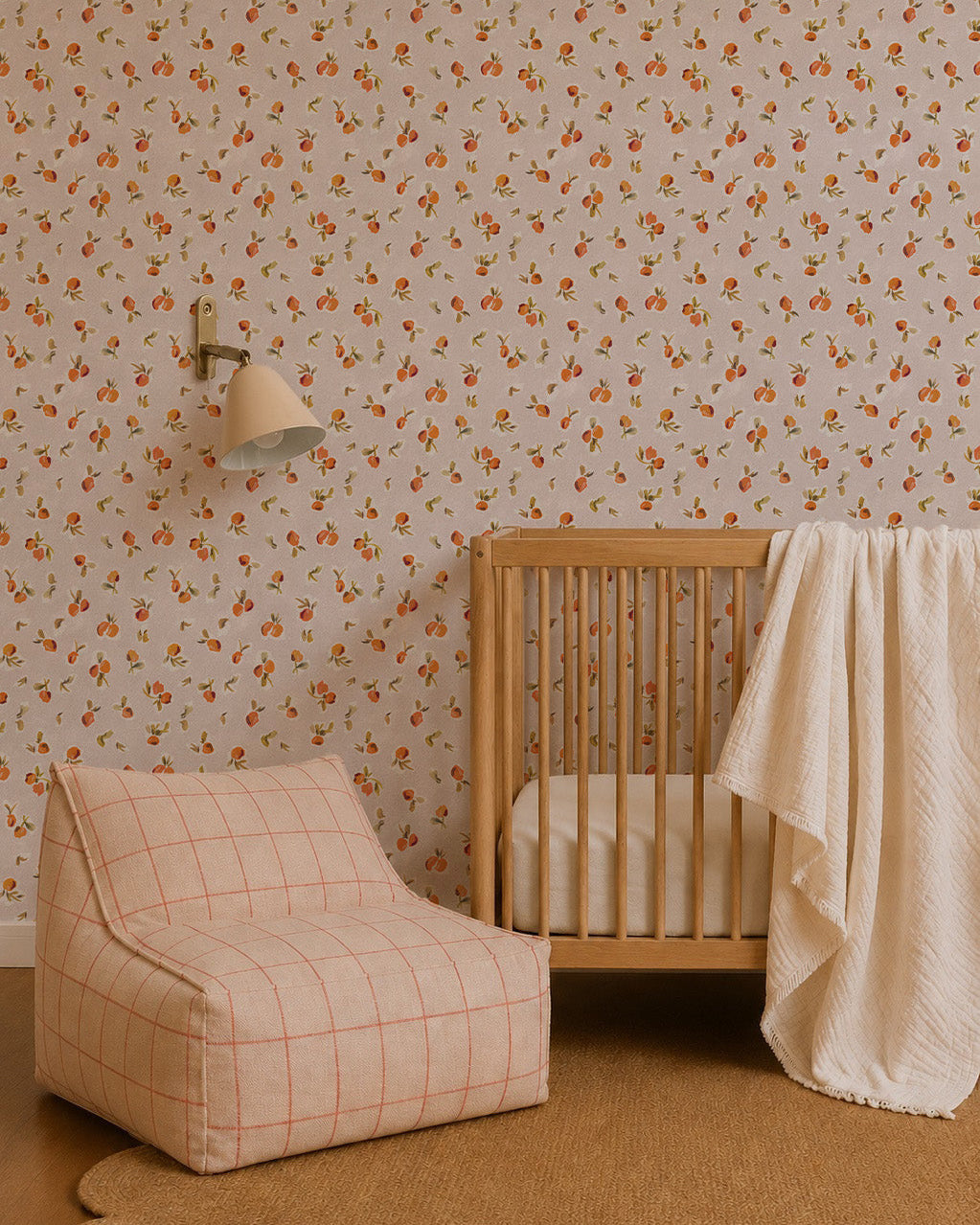 A cozy nursery with Ollies Oranges Wallpaper in pink, a wooden crib draped with a cream blanket, a modern beige and orange plaid lounge chair, a round woven rug, and a wall-mounted lamp.