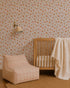 A cozy nursery with Ollies Oranges Wallpaper in pink, a wooden crib draped with a cream blanket, a modern beige and orange plaid lounge chair, a round woven rug, and a wall-mounted lamp.