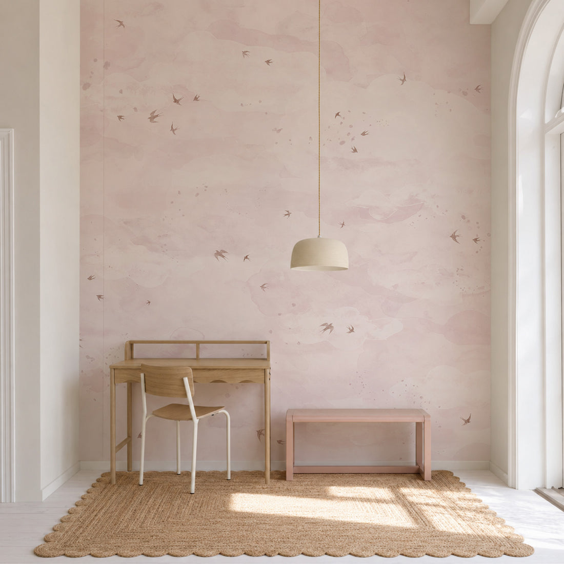 A room featuring an Opals Sky Birds Mural, Rose wall mural with watercolor-style clouds and small birds, styled with a wooden desk, a chair, and a low pink bench.