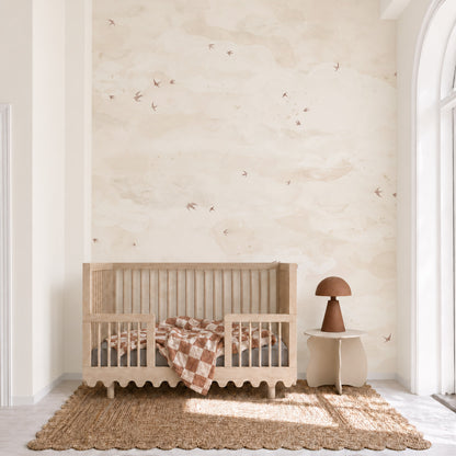 A wooden crib with a checkered blanket stands against a wall covered in Opals Sky Birds Mural, Sand wallpaper.