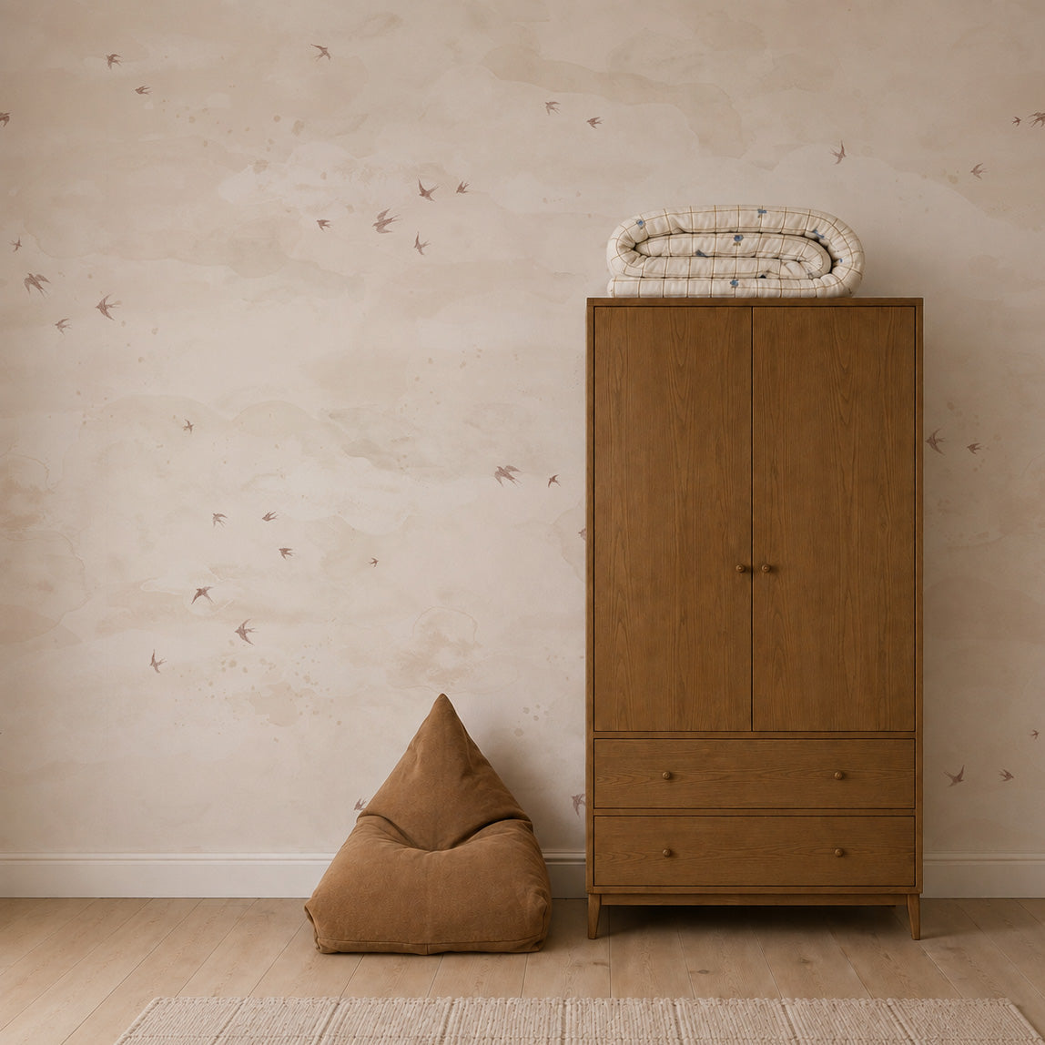 A nursery featuring Opals Sky Birds Mural, Sand, with small birds flying across a sandy-toned sky, a tall wooden wardrobe with a folded blanket on top, and a brown floor cushion.