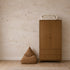 A nursery featuring Opals Sky Birds Mural, Sand, with small birds flying across a sandy-toned sky, a tall wooden wardrobe with a folded blanket on top, and a brown floor cushion.