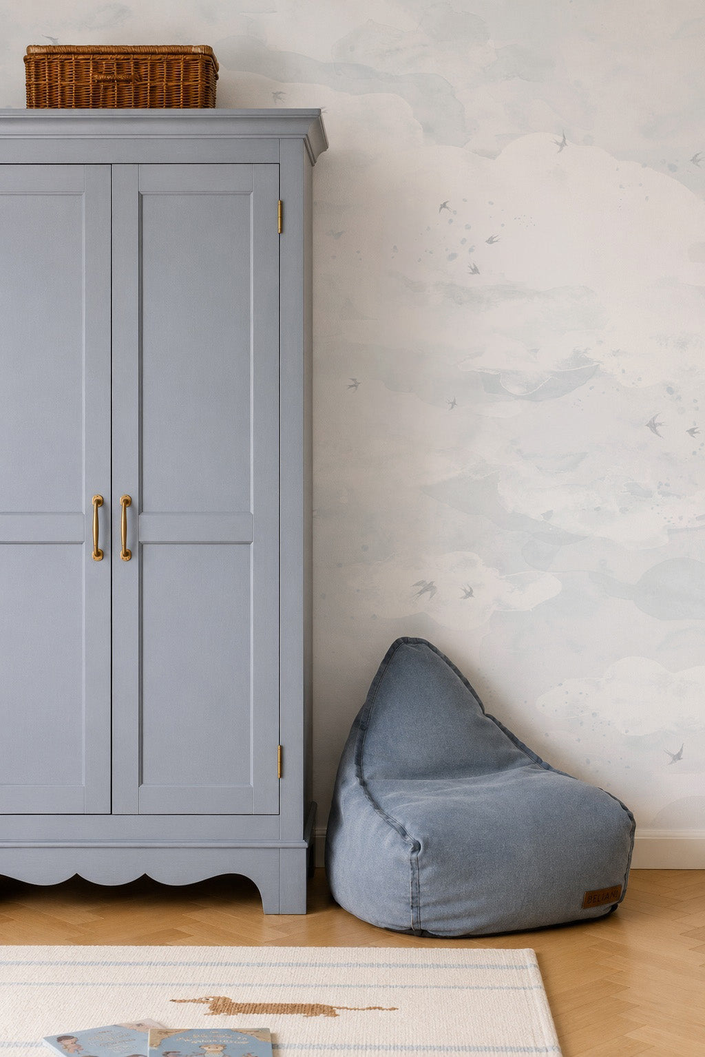 A grey wardrobe stands next to a blue beanbag chair against a wall covered in Opals Sky Birds Mural, Salt wallpaper featuring watercolor clouds.