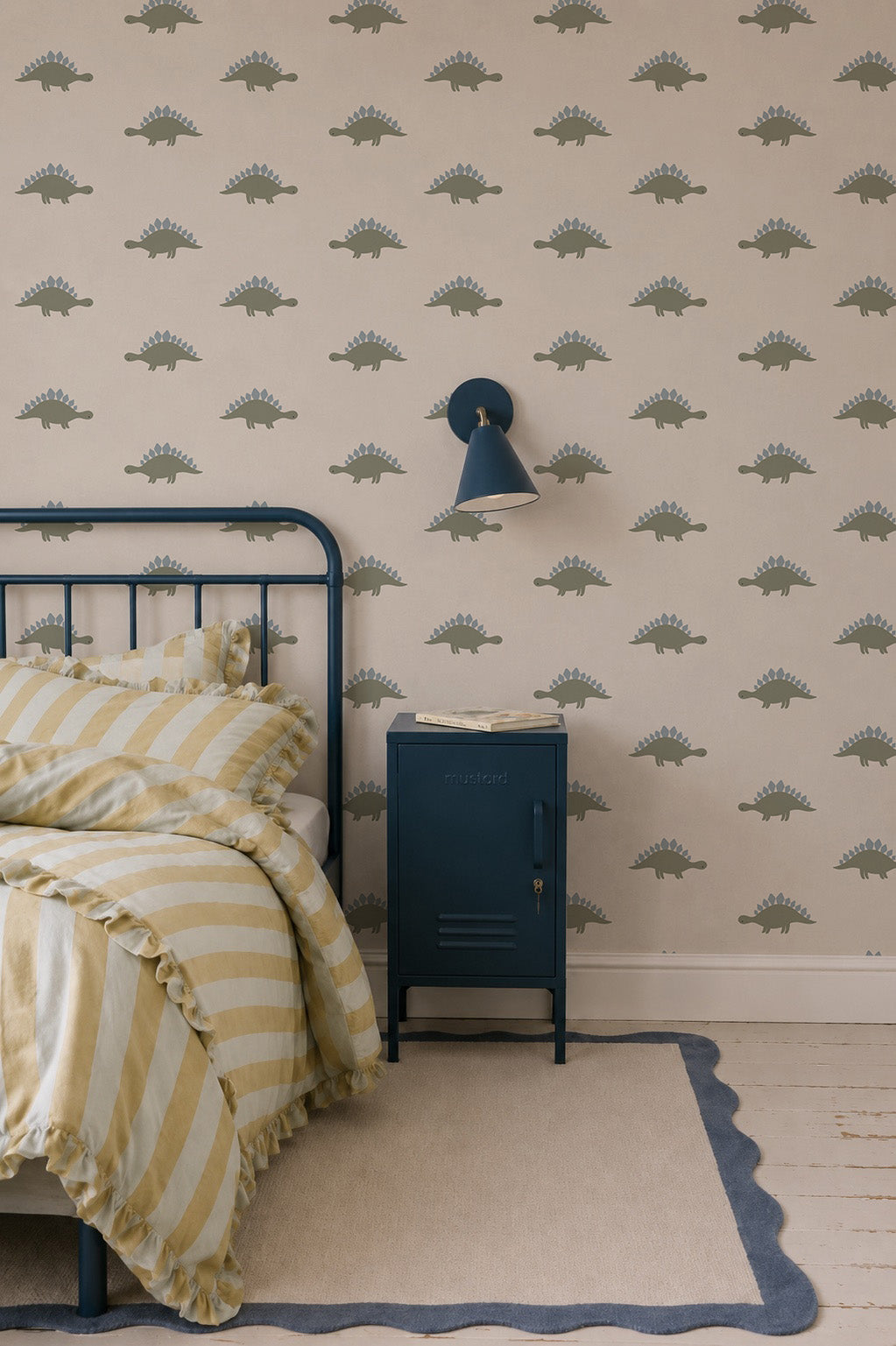 A cozy bedroom features a blue metal bed with yellow-striped bedding, a blue nightstand, a blue wall lamp, and Otis Dinosaur Wallpaper—perfect for playful home decor.