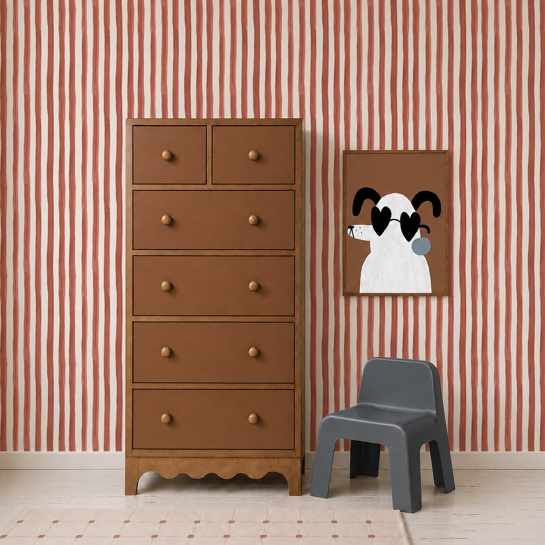 A brown dresser stands beside a small gray chair and a framed cartoon dog art on a wall adorned with Painted Stripe Wallpaper, Red, complemented by a beige carpet on the floor.