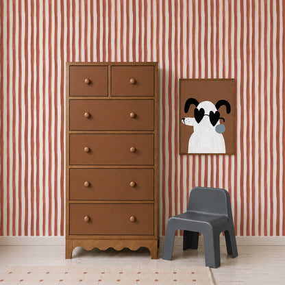 A brown dresser stands beside a small gray chair and a framed cartoon dog art on a wall adorned with Painted Stripe Wallpaper, Red, complemented by a beige carpet on the floor.