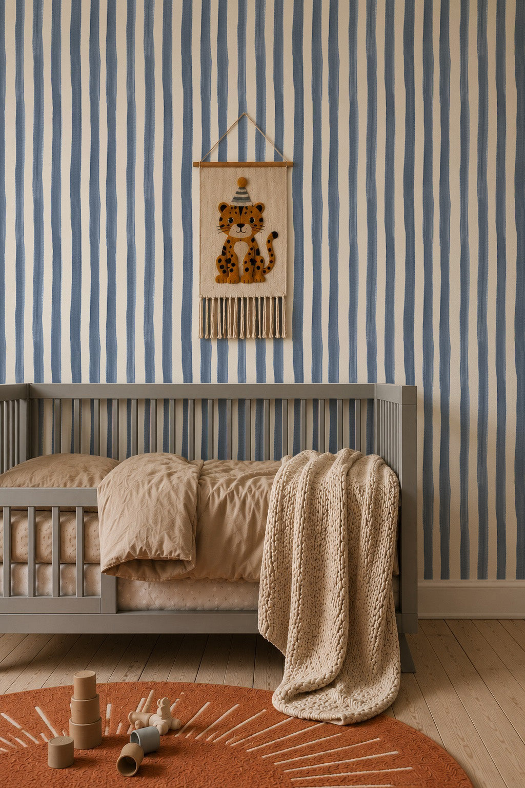 A cozy nursery with a gray crib, tan bedding, knitted beige blanket, rust-colored rug, and wooden toys. The wall features Painted Stripe Wallpaper in Blue and a hanging leopard decoration inspired by Stephanie Breeze.
