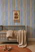 A cozy nursery with a gray crib, tan bedding, knitted beige blanket, rust-colored rug, and wooden toys. The wall features Painted Stripe Wallpaper in Blue and a hanging leopard decoration inspired by Stephanie Breeze.