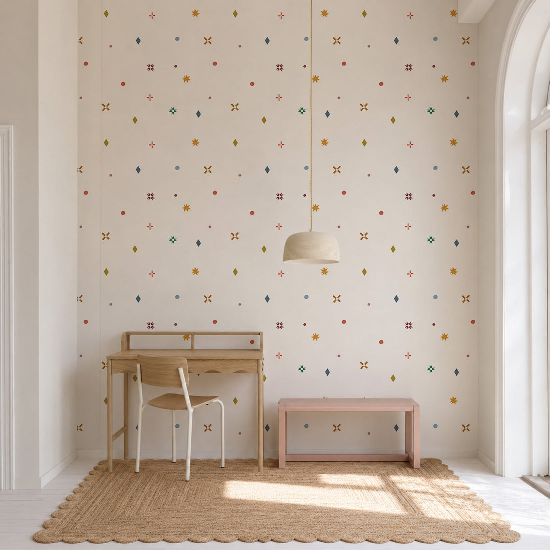 Minimalist room with a wooden desk, chair, pink bench, dome pendant light, and a woven rug. The wall features Pebble and Patch Wallpaper, Multi—a Scandinavian wallpaper with a colorful, small geometric pattern on a light background. Large arched window on the right.