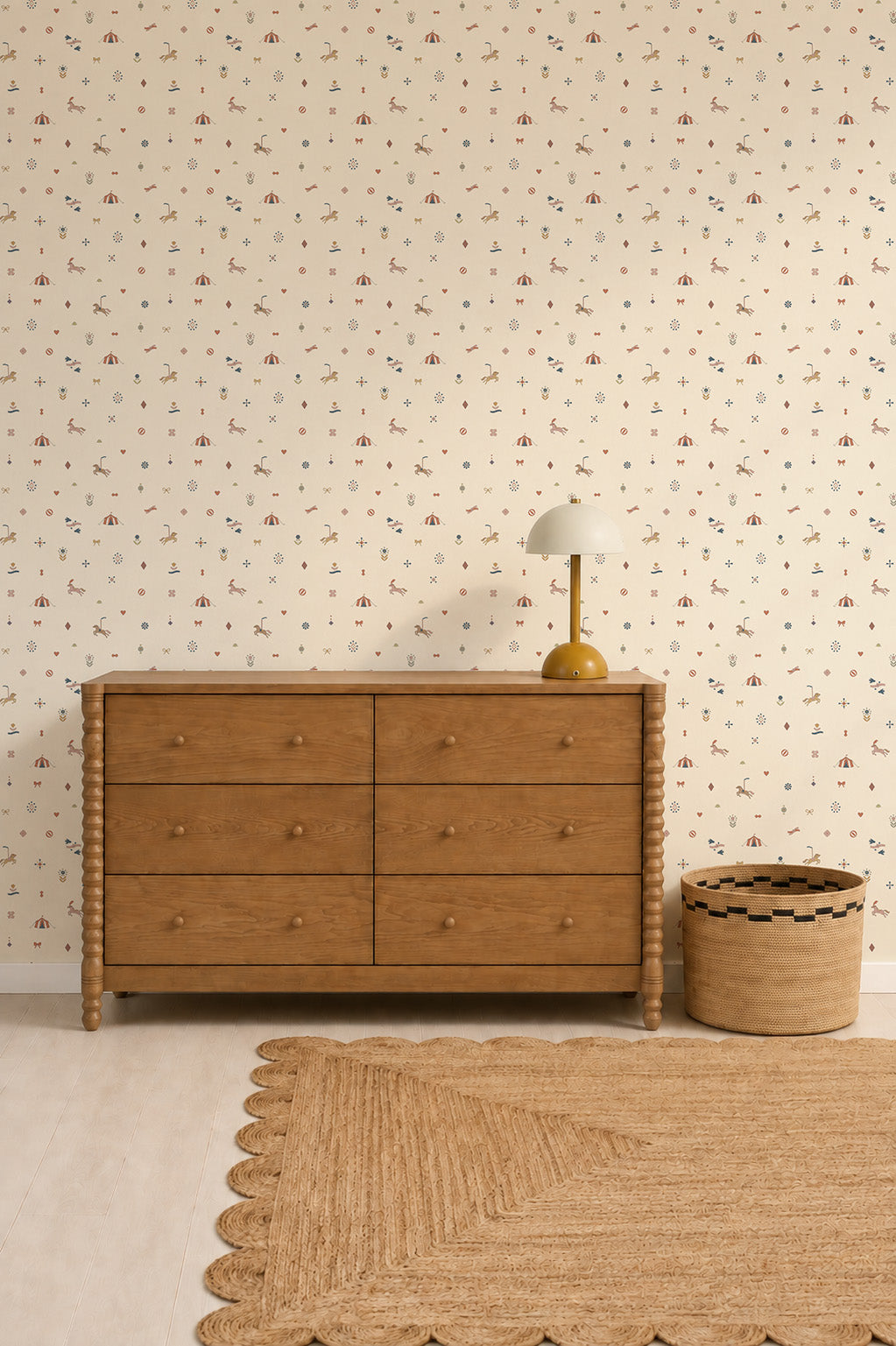 A wooden dresser with six drawers sits beside a woven basket and a lamp, set against a wall decorated with the charming Petite Circus Party Wallpaper and complemented by a tan rug with a scalloped edge on the light floor, creating the perfect touch of premium interior design.