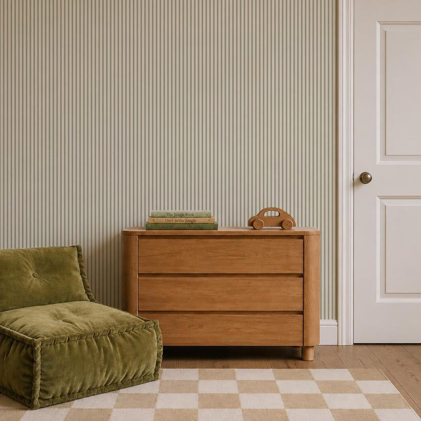 A wooden dresser with three drawers, two books, and a wooden toy on top sits next to a green cushioned chair. The wall is decorated with Pico Stripe Wallpaper in Green, and a door with a round knob is on the right. The floor features a checkered rug.