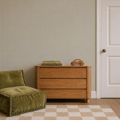 A wooden dresser with three drawers, two books, and a wooden toy on top sits next to a green cushioned chair. The wall is decorated with Pico Stripe Wallpaper in Green, and a door with a round knob is on the right. The floor features a checkered rug.