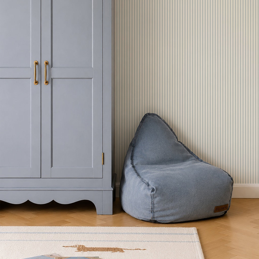 A light blue wardrobe with brass handles stands beside a blue bean bag chair on a wooden floor, set against calming Pico Stripe Wallpaper in Blue. A wicker basket is placed on top of the wardrobe.