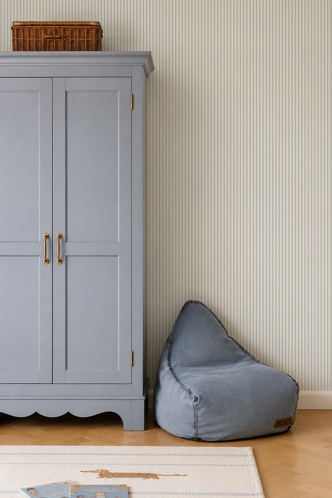 A light blue wardrobe with brass handles stands beside a blue bean bag chair on a wooden floor, set against calming Pico Stripe Wallpaper in Blue. A wicker basket is placed on top of the wardrobe.
