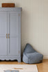 A light blue wardrobe with brass handles stands beside a blue bean bag chair on a wooden floor, set against calming Pico Stripe Wallpaper in Blue. A wicker basket is placed on top of the wardrobe.