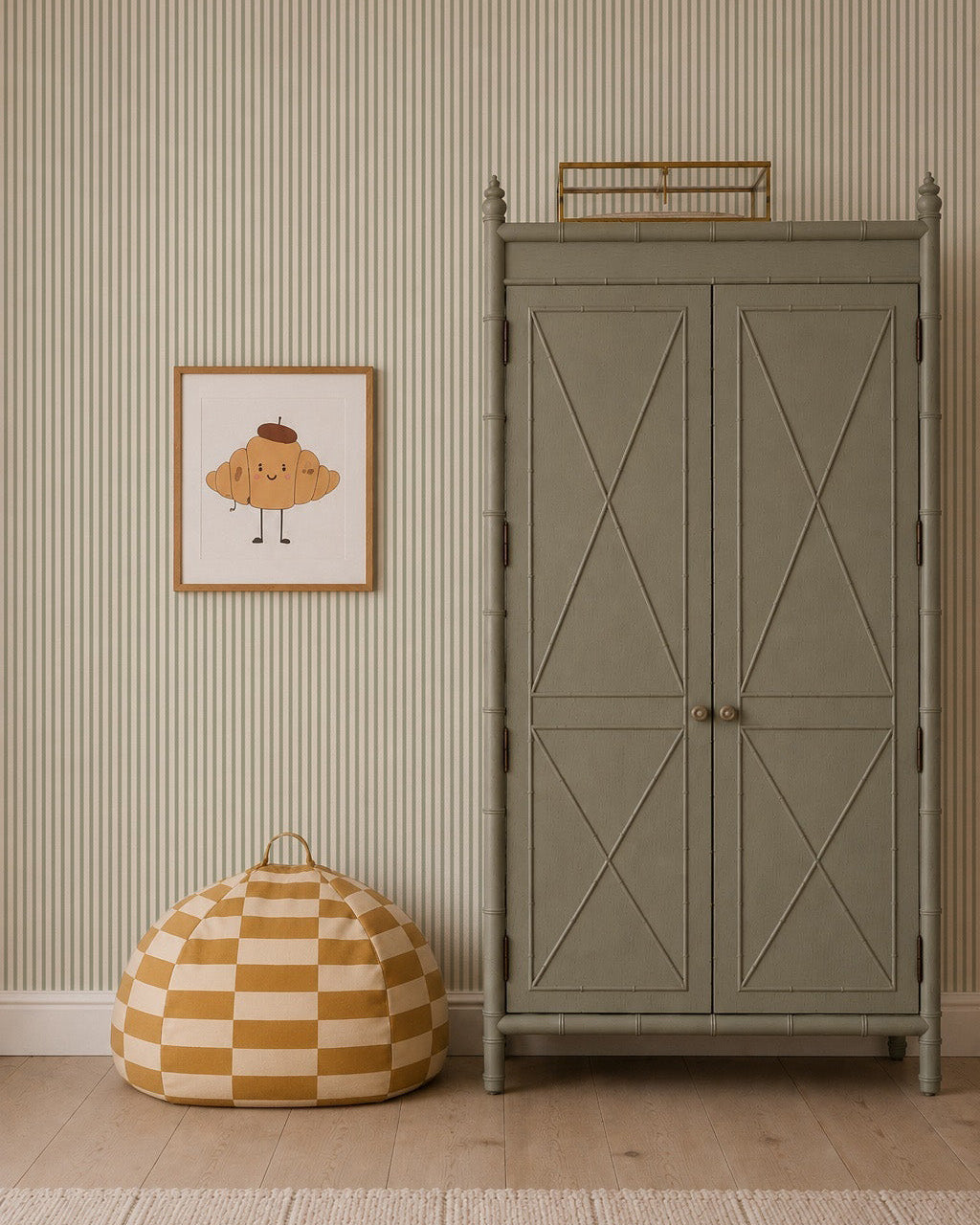 A sage green armoire stands against Pico Stripe Wallpaper in Green. To its left hangs a framed illustration of a smiling croissant, and below, a yellow-and-white checkered bean bag rests on the wooden floor.
