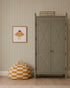 A sage green armoire stands against Pico Stripe Wallpaper in Green. To its left hangs a framed illustration of a smiling croissant, and below, a yellow-and-white checkered bean bag rests on the wooden floor.