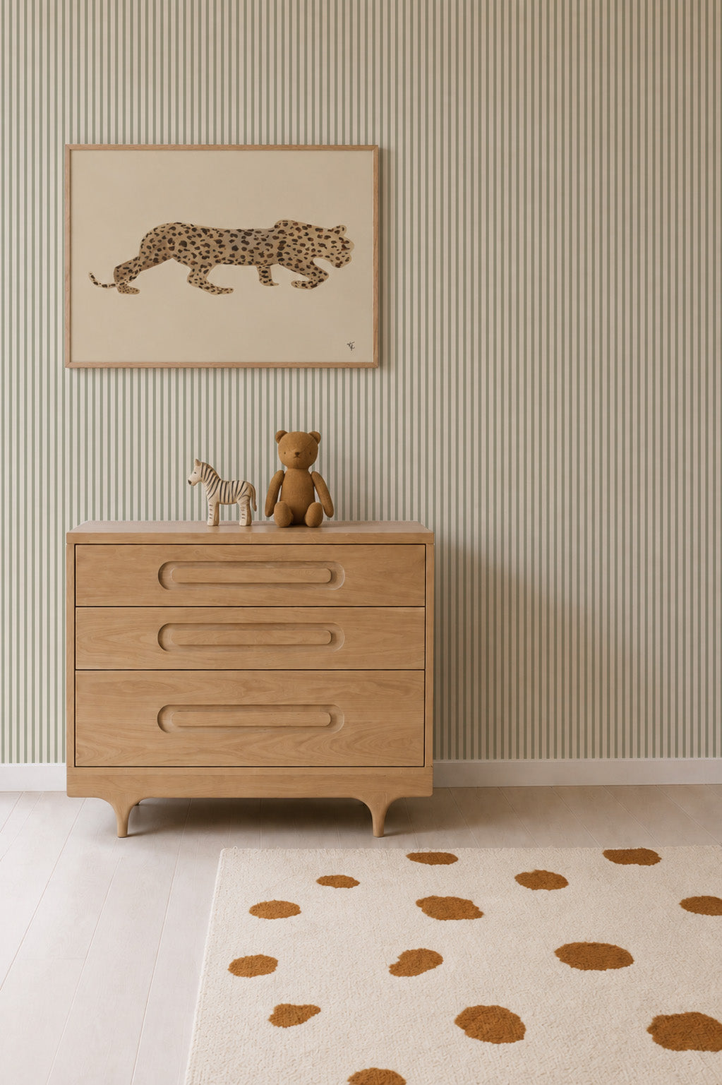 A wooden dresser with a teddy bear, zebra figurine, and leopard art above it sits against Pico Stripe Wallpaper in green, creating a charming and playful atmosphere. The nursery wallpaper ties the space together, complemented by a rug with brown spots.