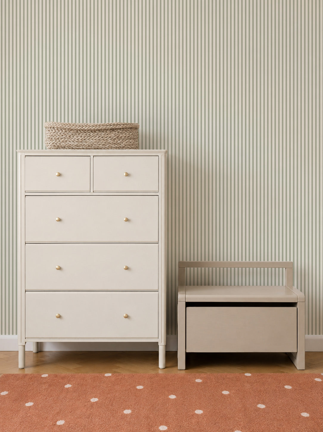 A white dresser with a woven basket on top stands next to a small bench with storage. The background features Pico Stripe Wallpaper in Green—perfect as nursery wallpaper—and an orange rug with white dots.