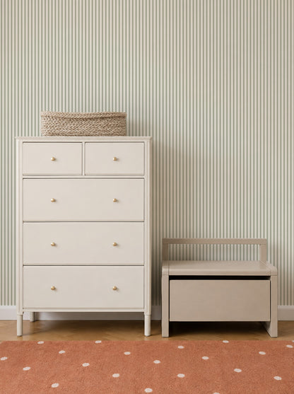 A white dresser with a woven basket on top stands next to a small bench with storage. The background features Pico Stripe Wallpaper in Green—perfect as nursery wallpaper—and an orange rug with white dots.