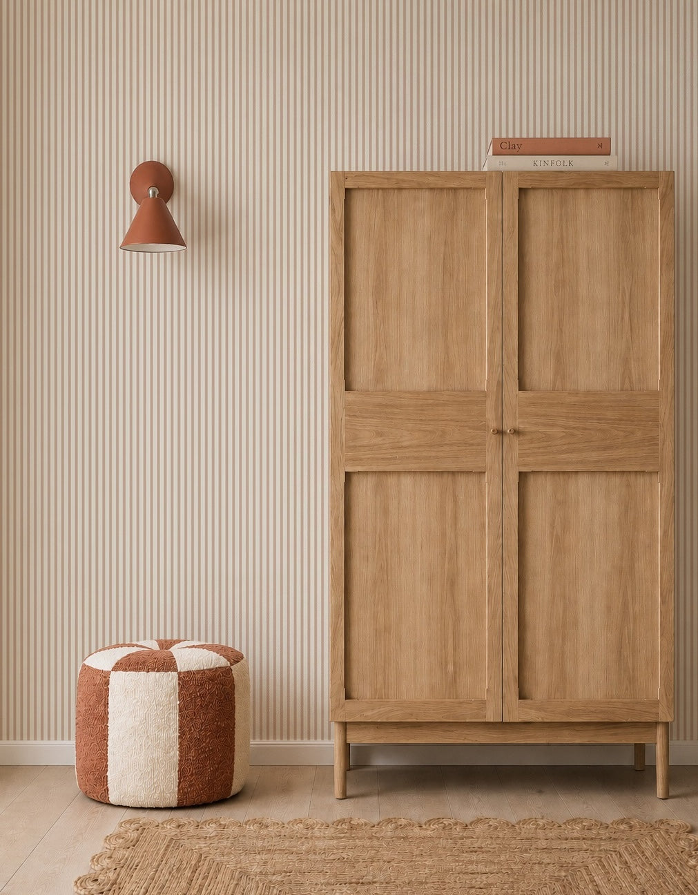 A minimalist room with a wooden wardrobe, a striped cream and rust pouf, woven rug, rust wall sconce, and Pico Stripe Wallpaper in Rose. Two books are stacked on the wardrobe.