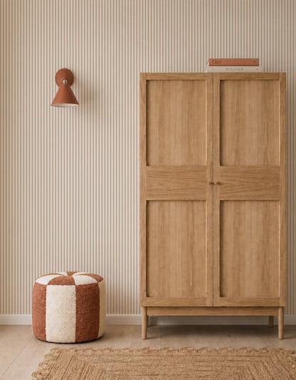A minimalist room with a wooden wardrobe, a striped cream and rust pouf, woven rug, rust wall sconce, and Pico Stripe Wallpaper in Rose. Two books are stacked on the wardrobe.