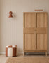 A minimalist room with a wooden wardrobe, a striped cream and rust pouf, woven rug, rust wall sconce, and Pico Stripe Wallpaper in Rose. Two books are stacked on the wardrobe.