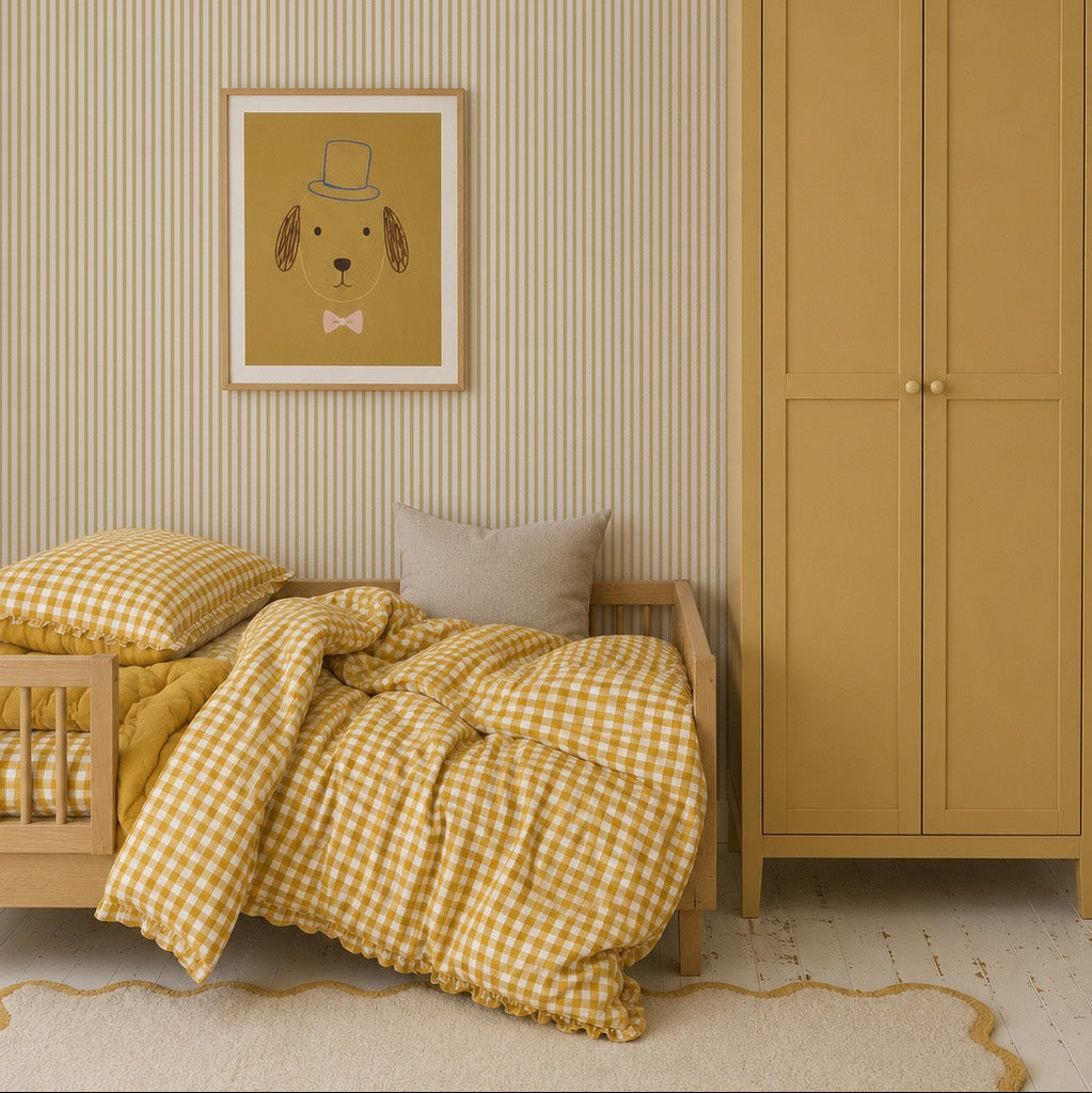 A cozy children’s bedroom features Pico Stripe Wallpaper in yellow, gingham bedding, a wooden bed and wardrobe, a beige cushion, a pendant light, and a framed dog-in-hat illustration.