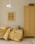 A cozy children’s bedroom features Pico Stripe Wallpaper in yellow, gingham bedding, a wooden bed and wardrobe, a beige cushion, a pendant light, and a framed dog-in-hat illustration.