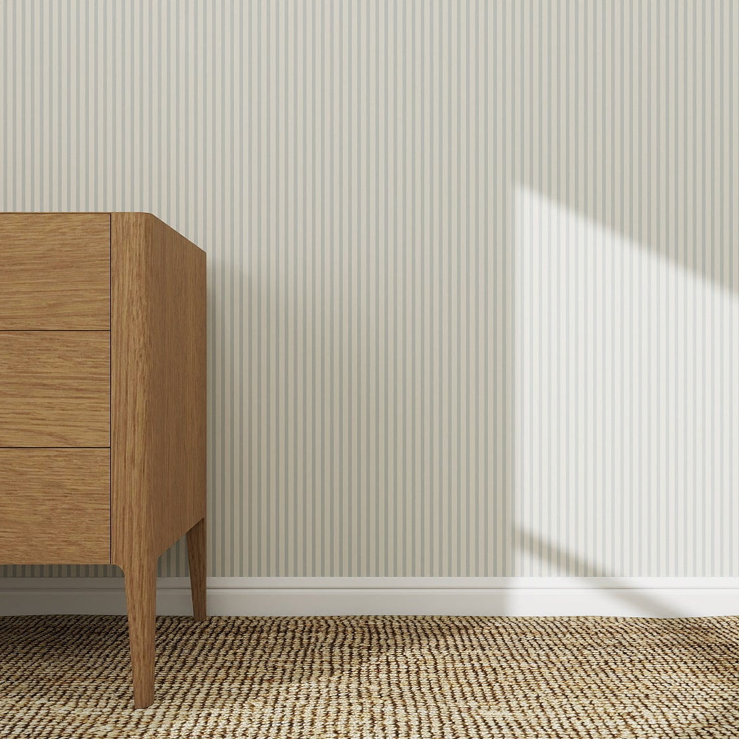 A wooden dresser stands on a woven rug against a light gray wall decorated with Pico Stripe Wallpaper Blue, featuring narrow vertical stripes. Sunlight casts gentle shadows, creating a bright and minimalistic interior scene.