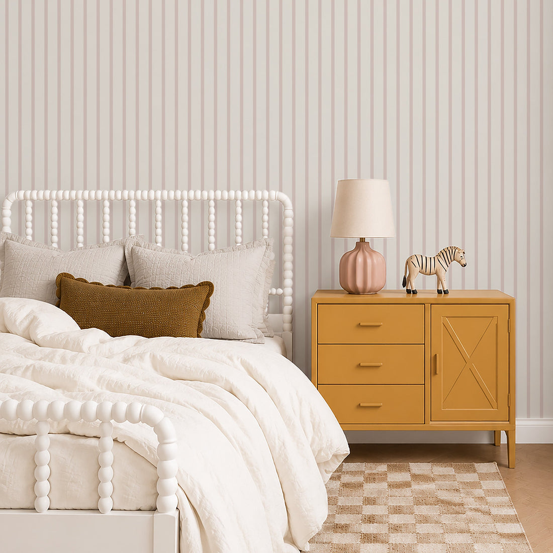 A cozy bedroom with a white spindle bed, cream bedding, and blush rose accents. A mustard yellow nightstand, lamp, ceramic vase, zebra figurine, and beige checkered rug sit on wood floors with Pinstripe Wallpaper in Blush Rose.