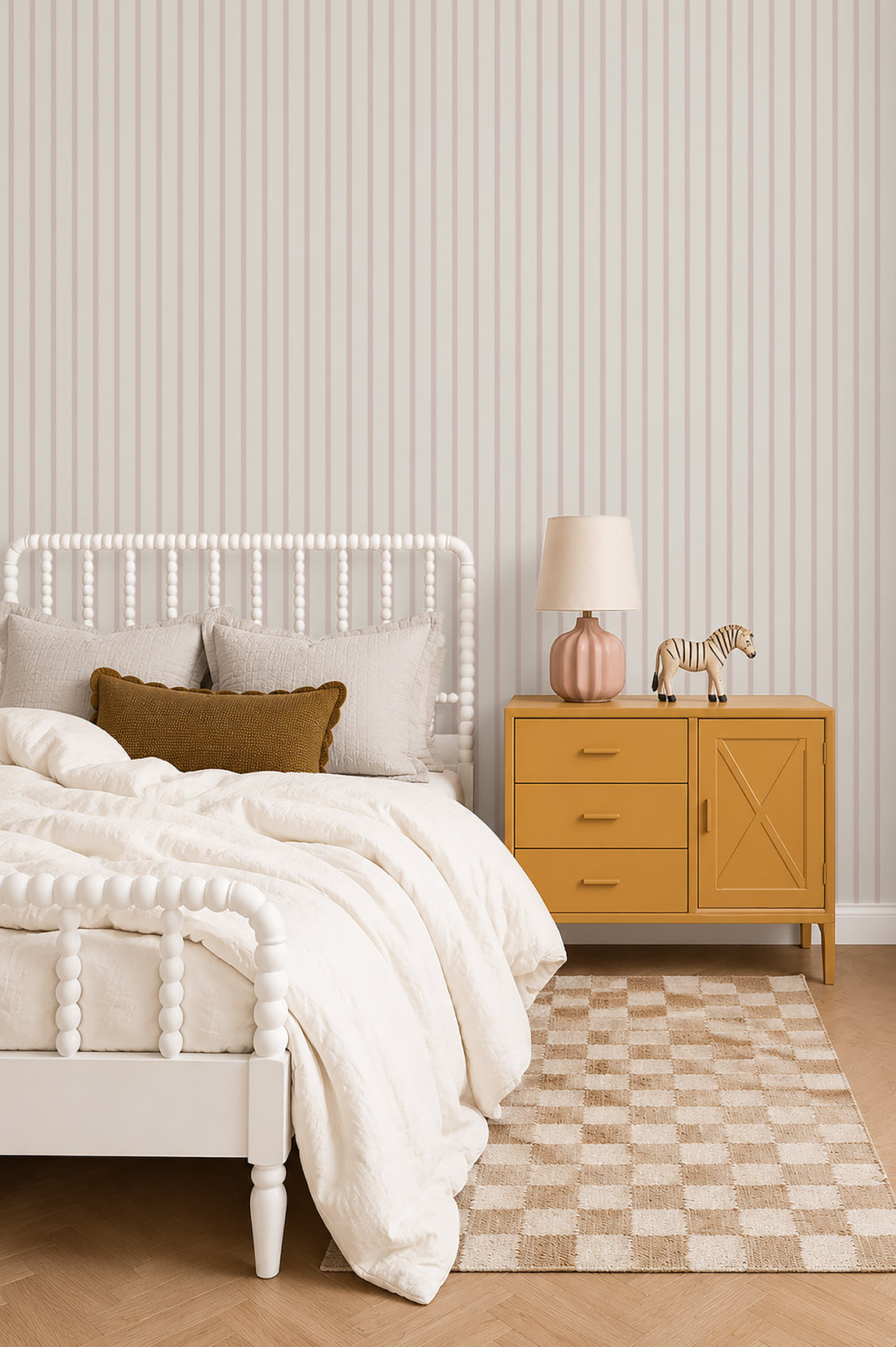 A cozy bedroom with a white spindle bed, cream bedding, and blush rose accents. A mustard yellow nightstand, lamp, ceramic vase, zebra figurine, and beige checkered rug sit on wood floors with Pinstripe Wallpaper in Blush Rose.
