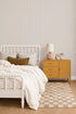 A cozy bedroom with a white spindle bed, cream bedding, and blush rose accents. A mustard yellow nightstand, lamp, ceramic vase, zebra figurine, and beige checkered rug sit on wood floors with Pinstripe Wallpaper in Blush Rose.