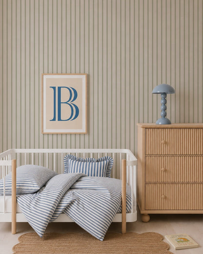 A nursery with a white crib, blue and white striped bedding, Pinstripe Wallpaper Green, a chest of drawers with a blue lamp, a framed letter B on the wall, a round jute rug, and a children&